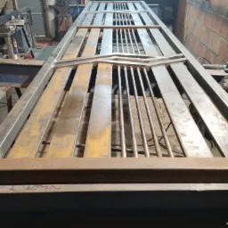 Metalowa konstrukcja bramy wjazdowej w trakcie spawania, widoczne elementy ozdobne i pręty, na tle warsztatu z narzędziami i ceglaną ścianą.