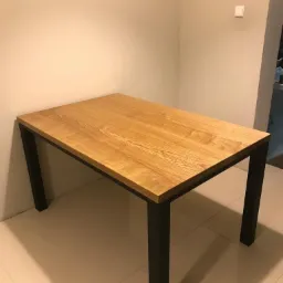 Stół z blatem z jasnego drewna o wyraźnym usłojeniu i prostych, czarnych, metalowych nogach, ustawiony na podłodze z jasnych płytek ceramicznych w rogu pokoju.