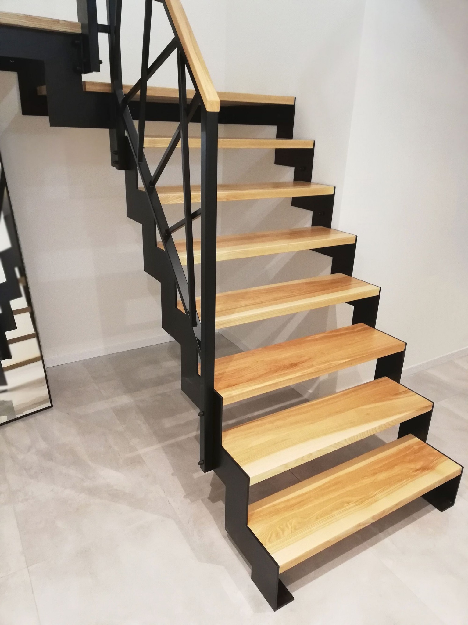 Nowoczesne schody z drewnianymi stopniami i czarną metalową konstrukcją, minimalistyczny design, jasna podłoga i ściana w tle, ujęcie z dołu.
