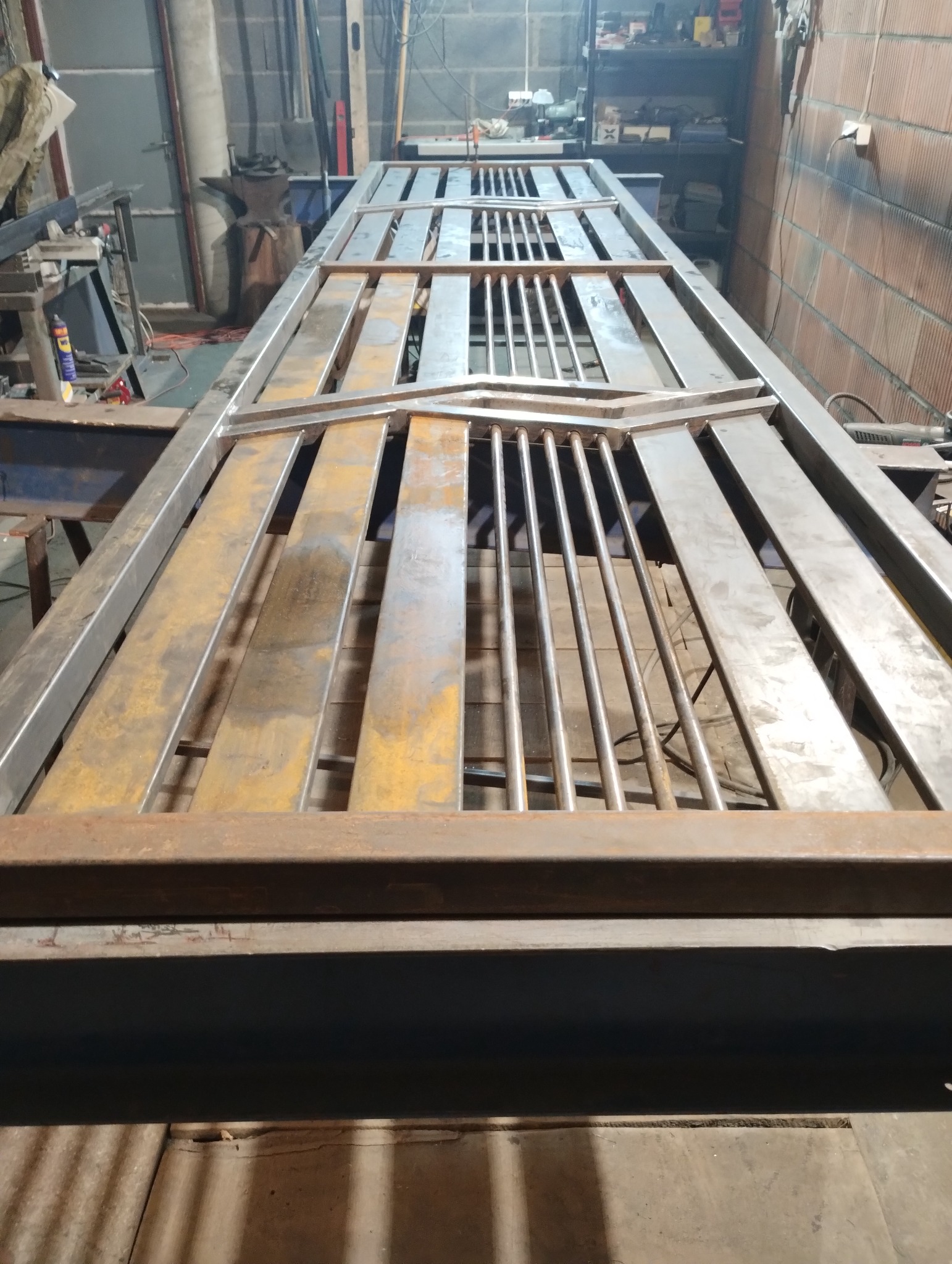 Metalowa konstrukcja bramy wjazdowej w trakcie spawania, widoczne elementy ozdobne i pręty, na tle warsztatu z narzędziami i ceglaną ścianą.