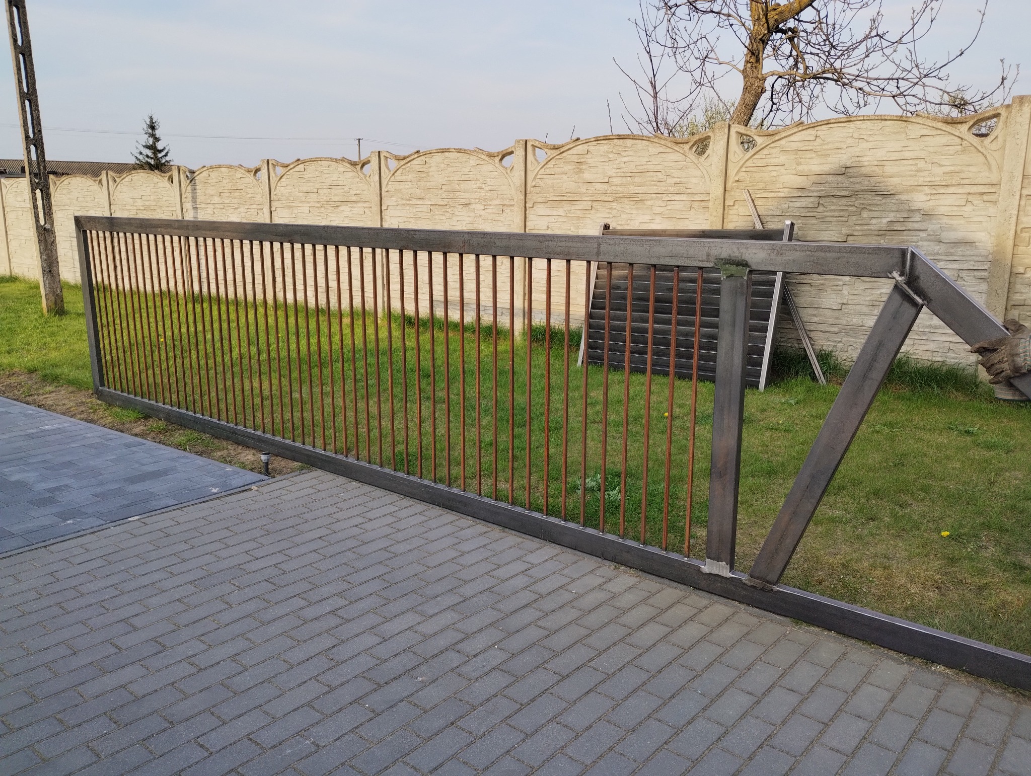 Surowa, metalowa konstrukcja przesuwnej bramy wjazdowej z pionowymi prętami, widoczna na tle betonowego ogrodzenia i kostki brukowej.