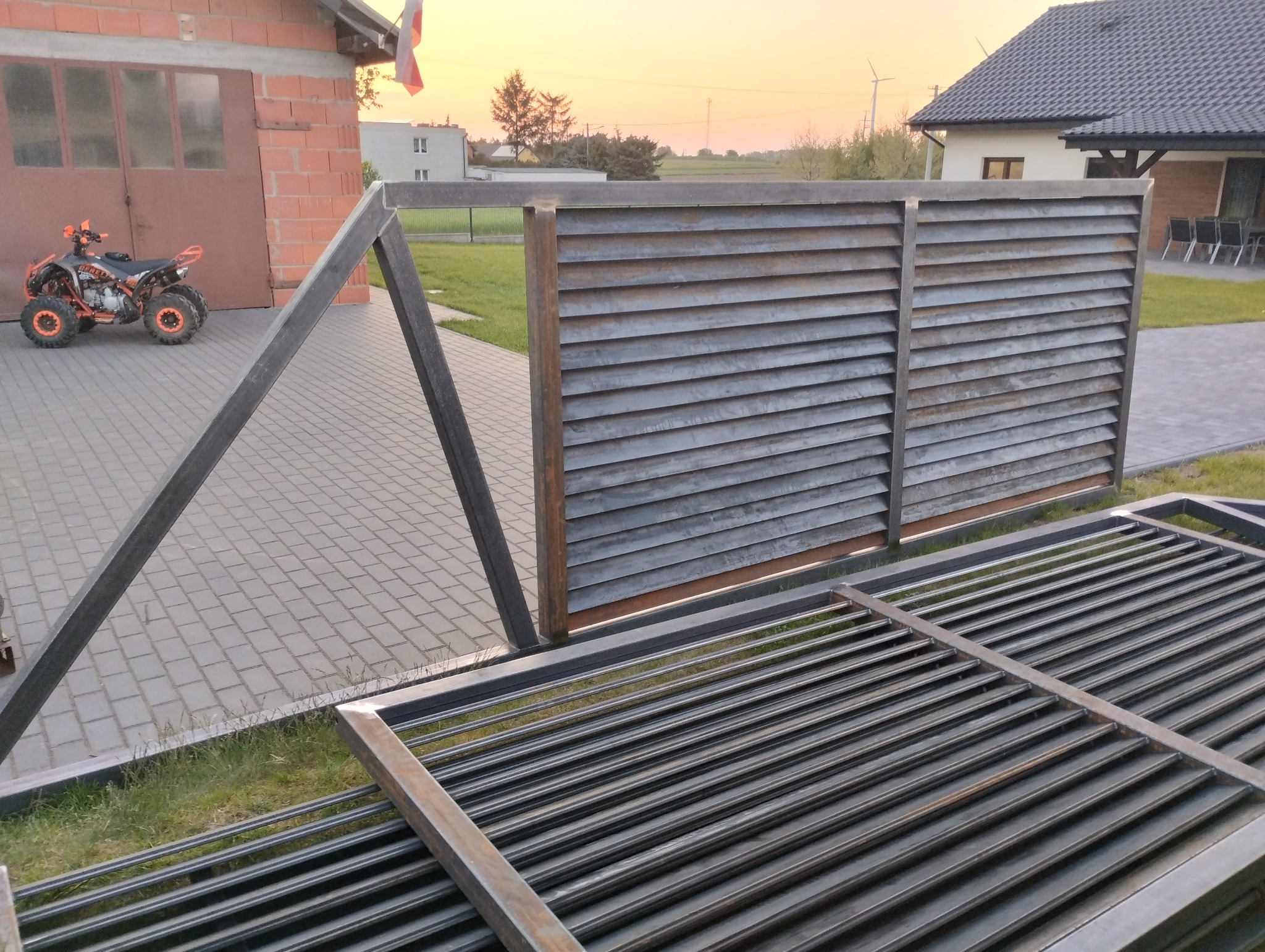 Fragment częściowo zmontowanej, metalowej bramy wjazdowej z poziomymi lamelami, widocznej na tle posesji w Kramsku, obok stoi pomarańczowy quad.