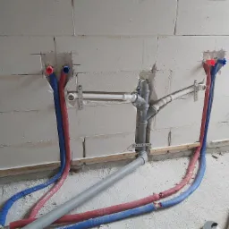 Wewnętrzna instalacja wodno-kanalizacyjna w trakcie montażu, widoczne rury PEX w izolacji czerwonej i niebieskiej, oraz szare rury kanalizacyjne z kształtkami, zamocowane do ściany z betonu...