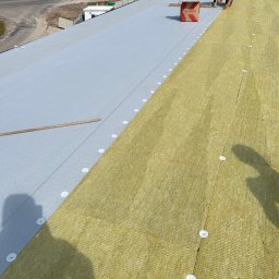 Term- Bud Łukasz Osmólski - Montaż izolacji termicznej z wełny mineralnej na płaskim dachu, widoczna warstwa hydroizolacyjna, pracownik z narzędziami, paczka wełny mineralnej Klimas, otoczenie zieleni i droga.