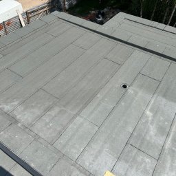 Term- Bud Łukasz Osmólski - Płaski dach pokryty szarą papą bitumiczną z widocznym odpływem wody, fragmentem ogrodzenia i białym samochodem dostawczym w tle.