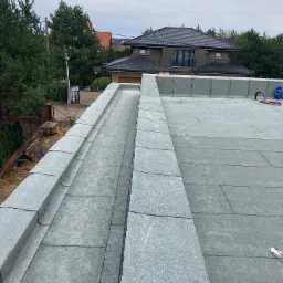 Widok z góry na płaski dach pokryty papą termozgrzewalną z widoczną attyką i systemem odprowadzania wody, w tle dom jednorodzinny i drzewa.