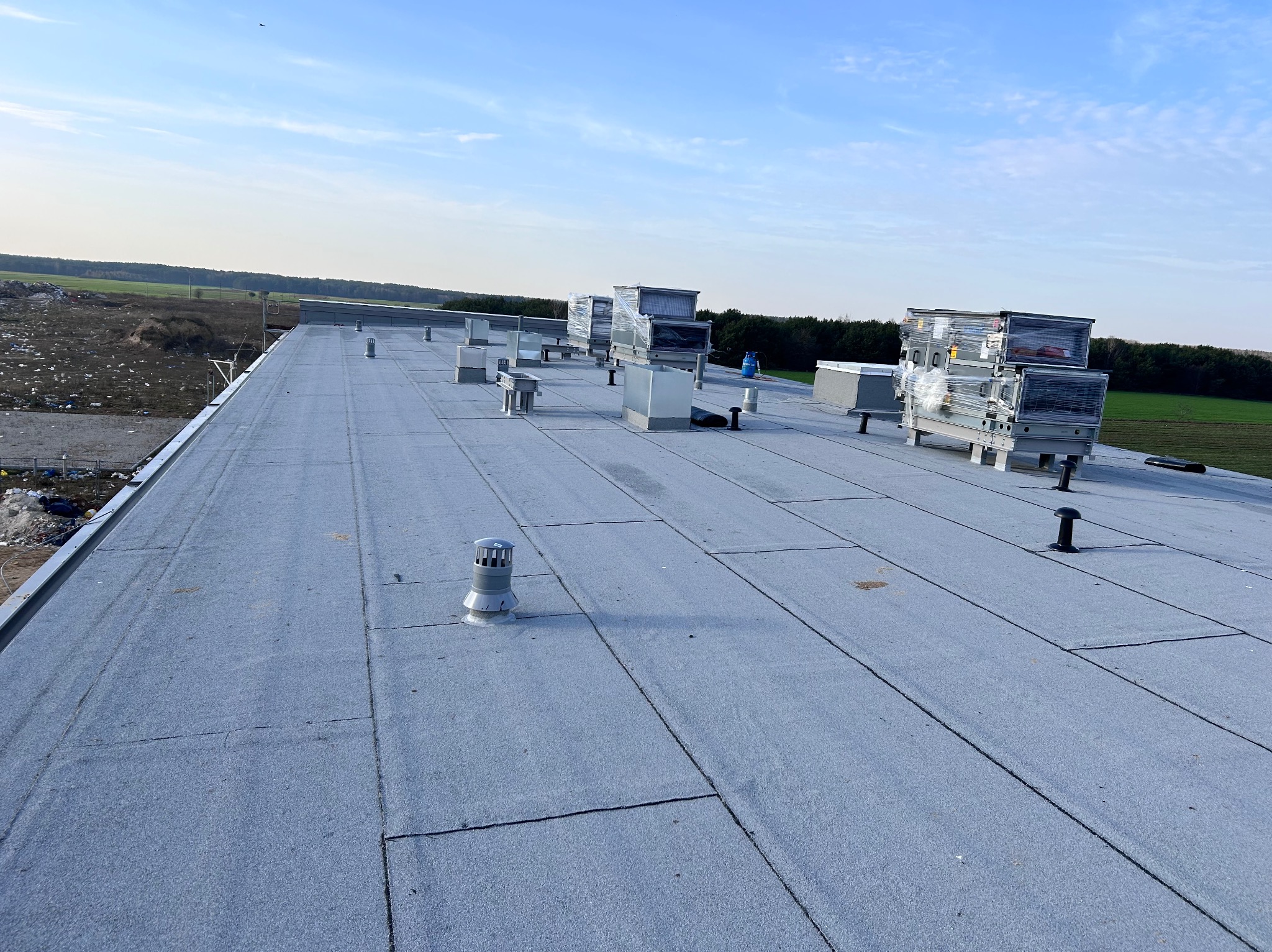 Płaski dach pokryty papą termozgrzewalną z widocznymi kominkami wentylacyjnymi i centralami klimatyzacyjnymi w folii ochronnej, na tle zieleni i błękitnego nieba.