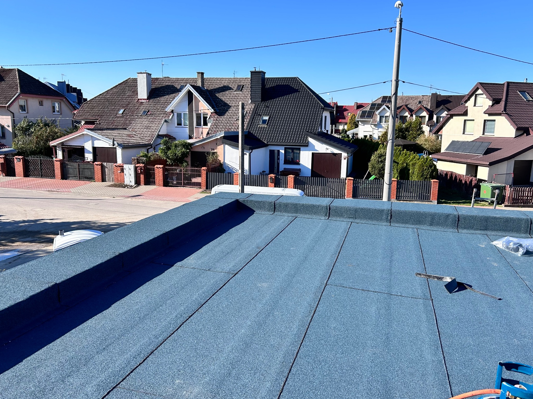 Niebieski dach płaski pokryty papą termozgrzewalną, widoczne narzędzie dekarskie i butla gazowa, w tle zabudowa jednorodzinna w słoneczny dzień.