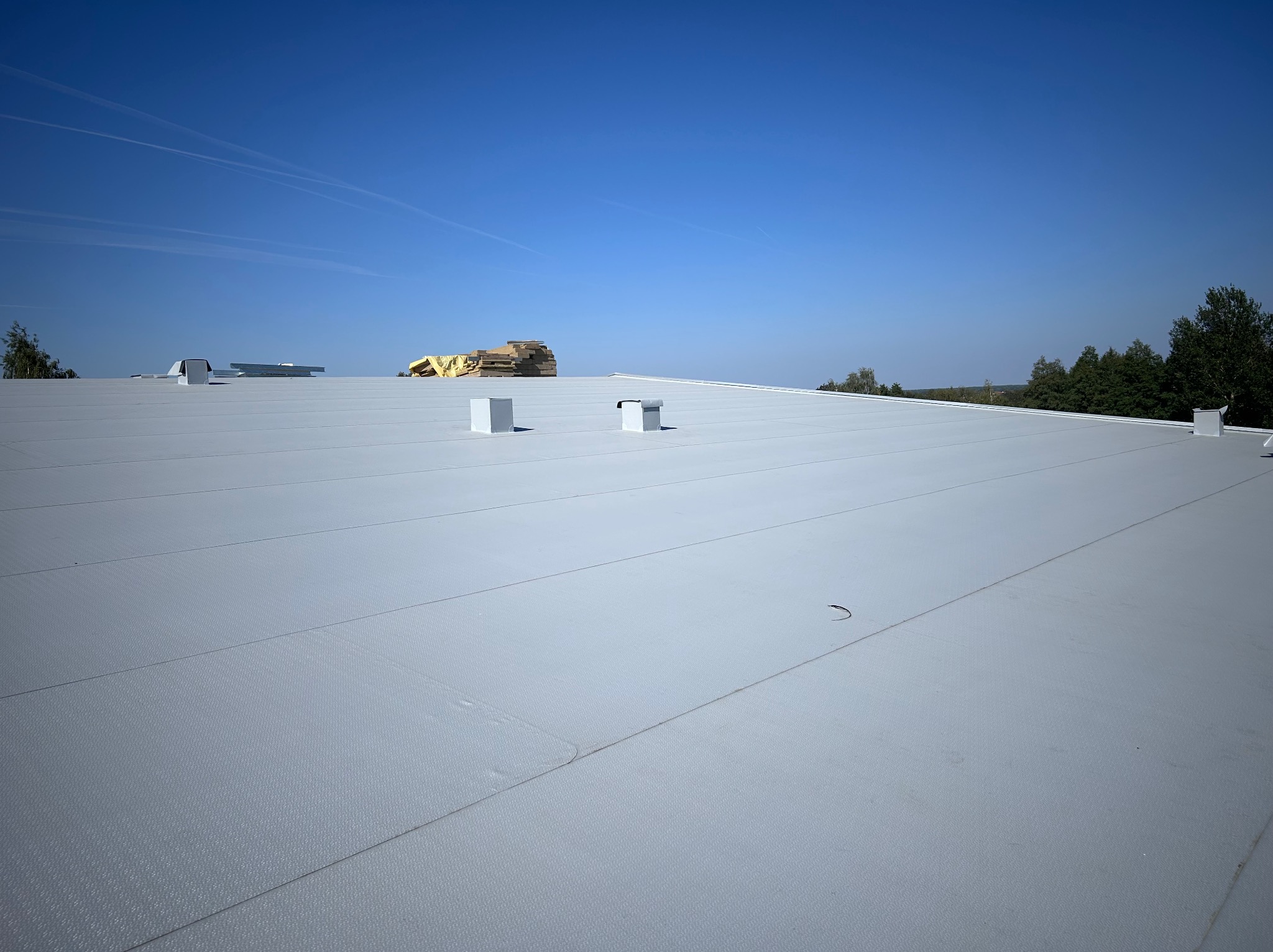 Jasnoszary, płaski dach pokryty membraną PVC, z widocznymi kominkami wentylacyjnymi i stertą materiałów budowlanych w oddali, na tle błękitnego nieba.