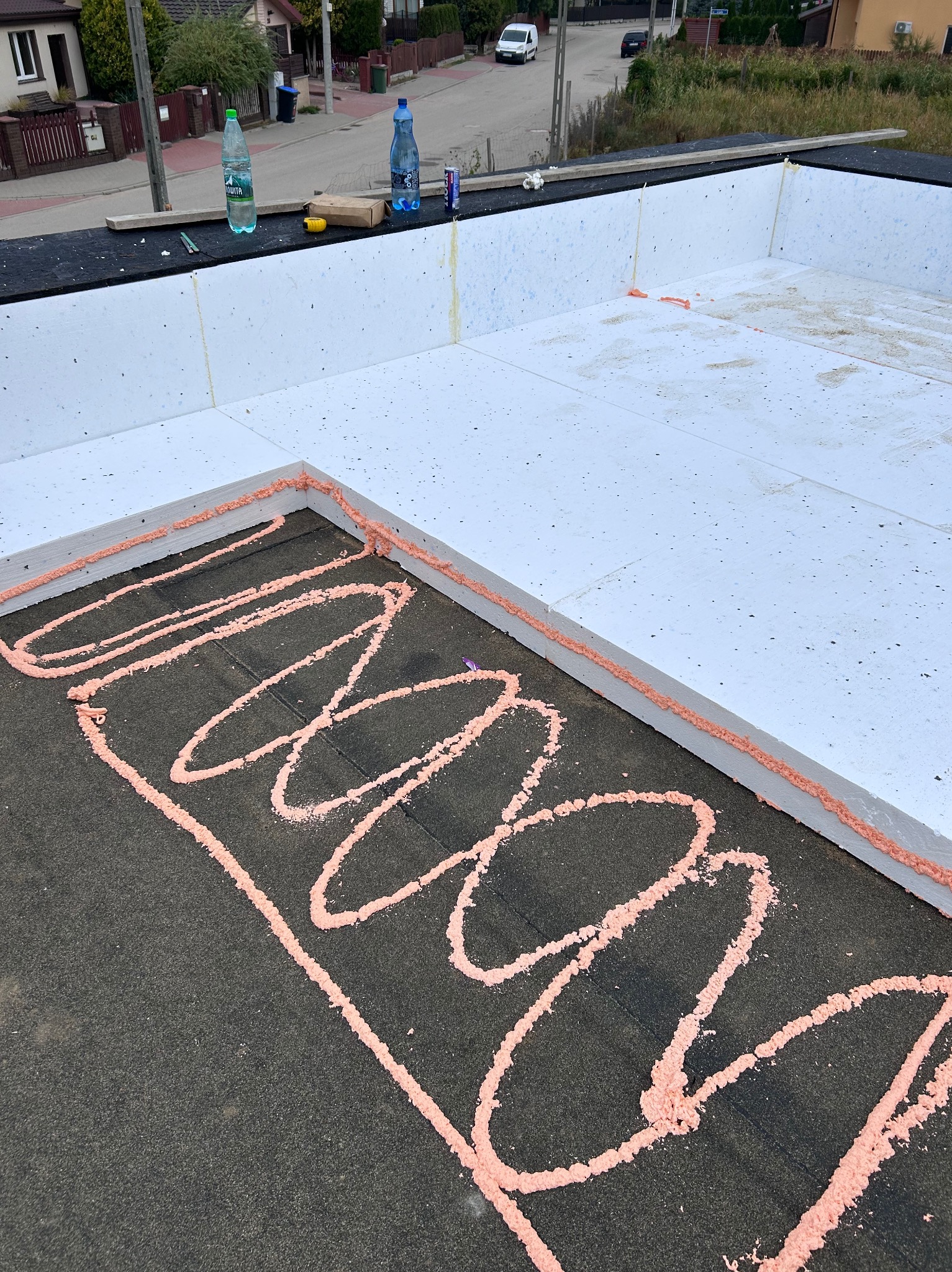 Ocieplenie dachu styropianem z użyciem pianki montażowej, widok z góry na płaski dach w trakcie prac izolacyjnych, butelki wody w tle.