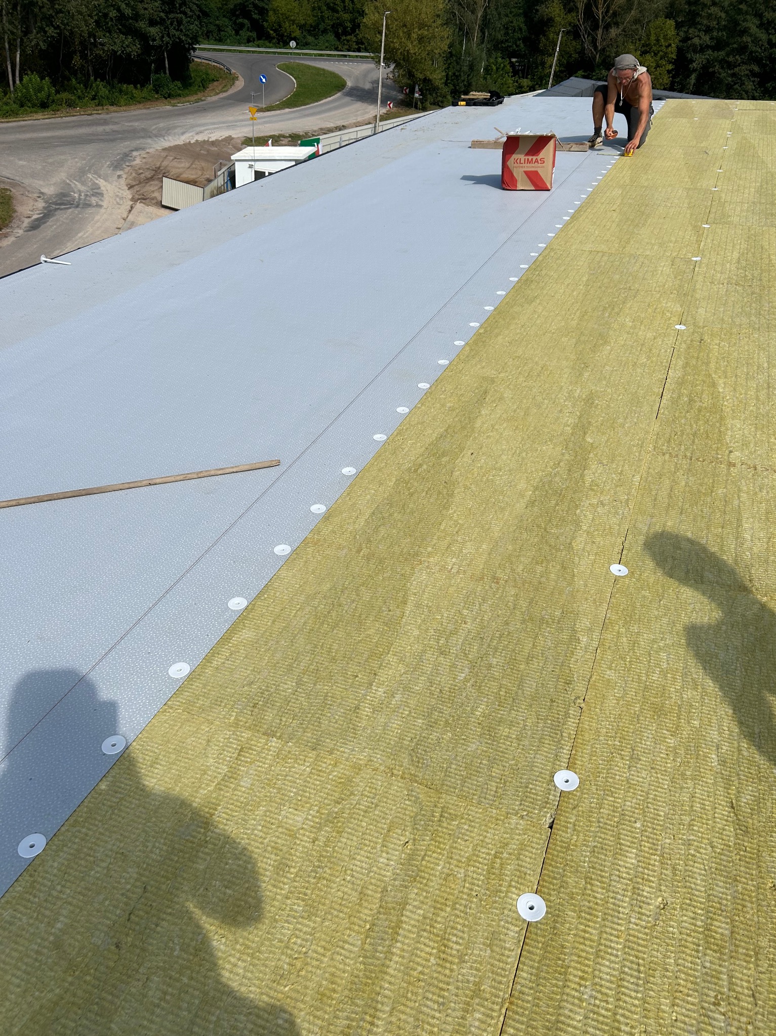 Montaż izolacji termicznej z wełny mineralnej na płaskim dachu, widoczna warstwa hydroizolacyjna, pracownik z narzędziami, paczka wełny mineralnej Klimas, otoczenie zieleni i droga.