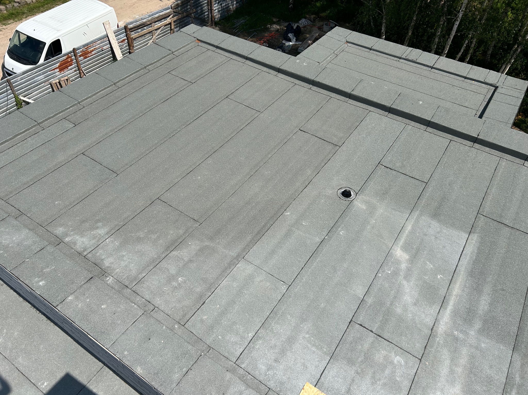 Płaski dach pokryty szarą papą bitumiczną z widocznym odpływem wody, fragmentem ogrodzenia i białym samochodem dostawczym w tle.