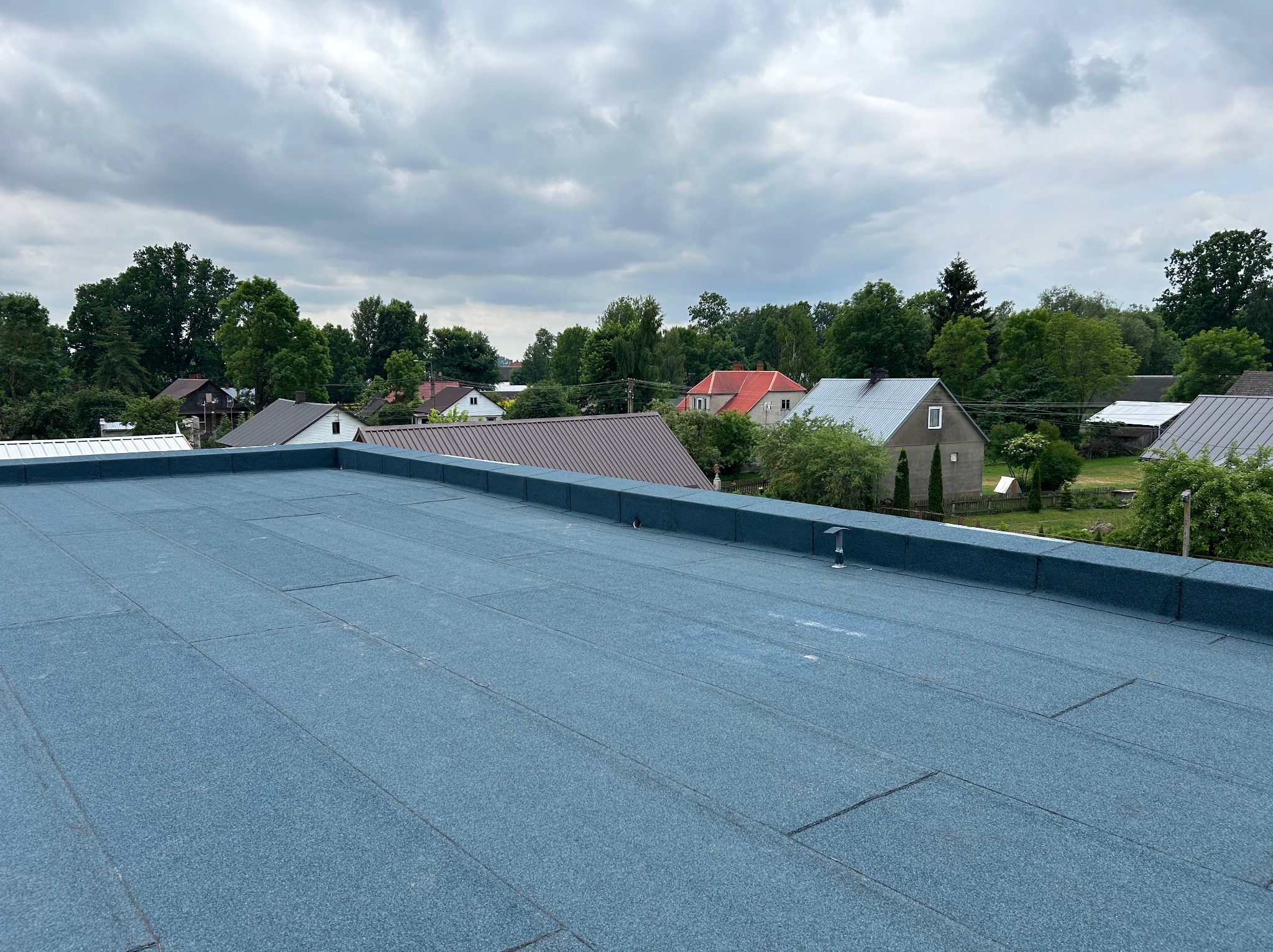 Niebieski, płaski dach pokryty papą na tle zabudowań jednorodzinnych i pochmurnego nieba.