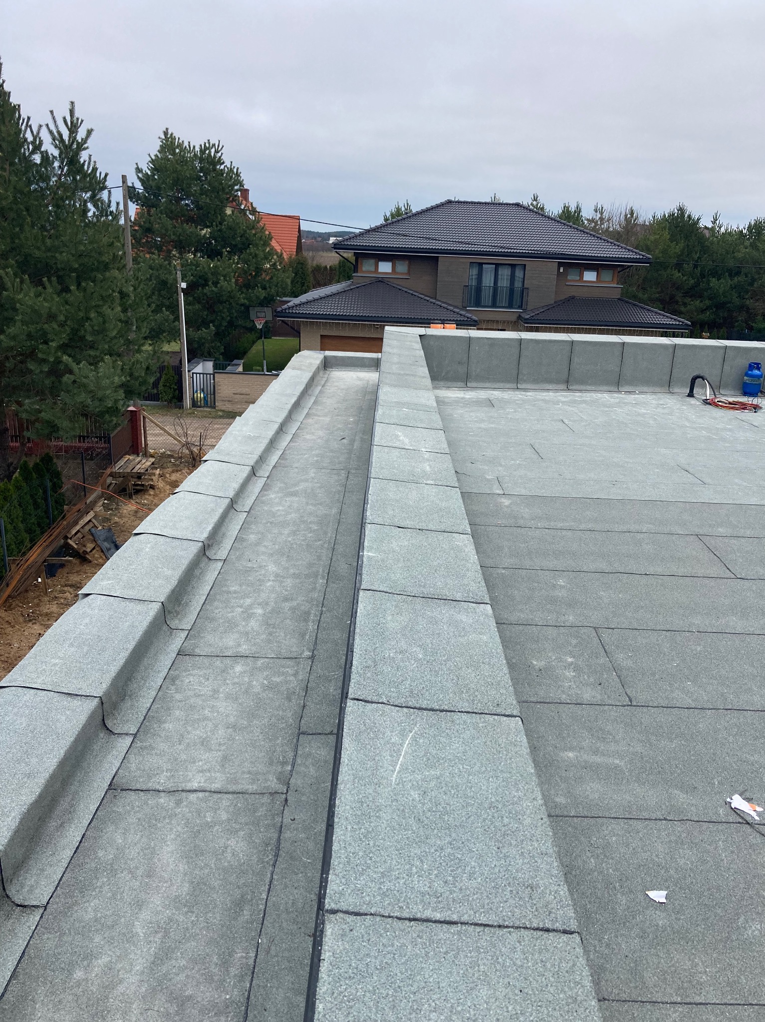 Widok z góry na płaski dach pokryty papą termozgrzewalną z widoczną attyką i systemem odprowadzania wody, w tle dom jednorodzinny i drzewa.