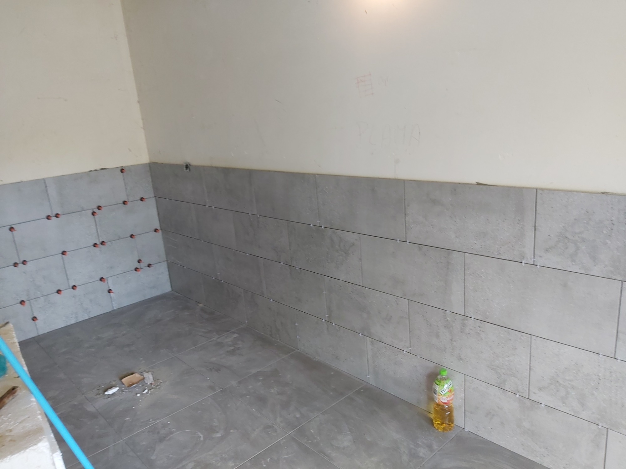 Świeżo ułożone szare płytki na ścianie i podłodze z użyciem czerwonych krzyżyków dystansowych, butelka oleju stoi w rogu, ślady po zaprawie na podłodze, surowy stan wykończenia.