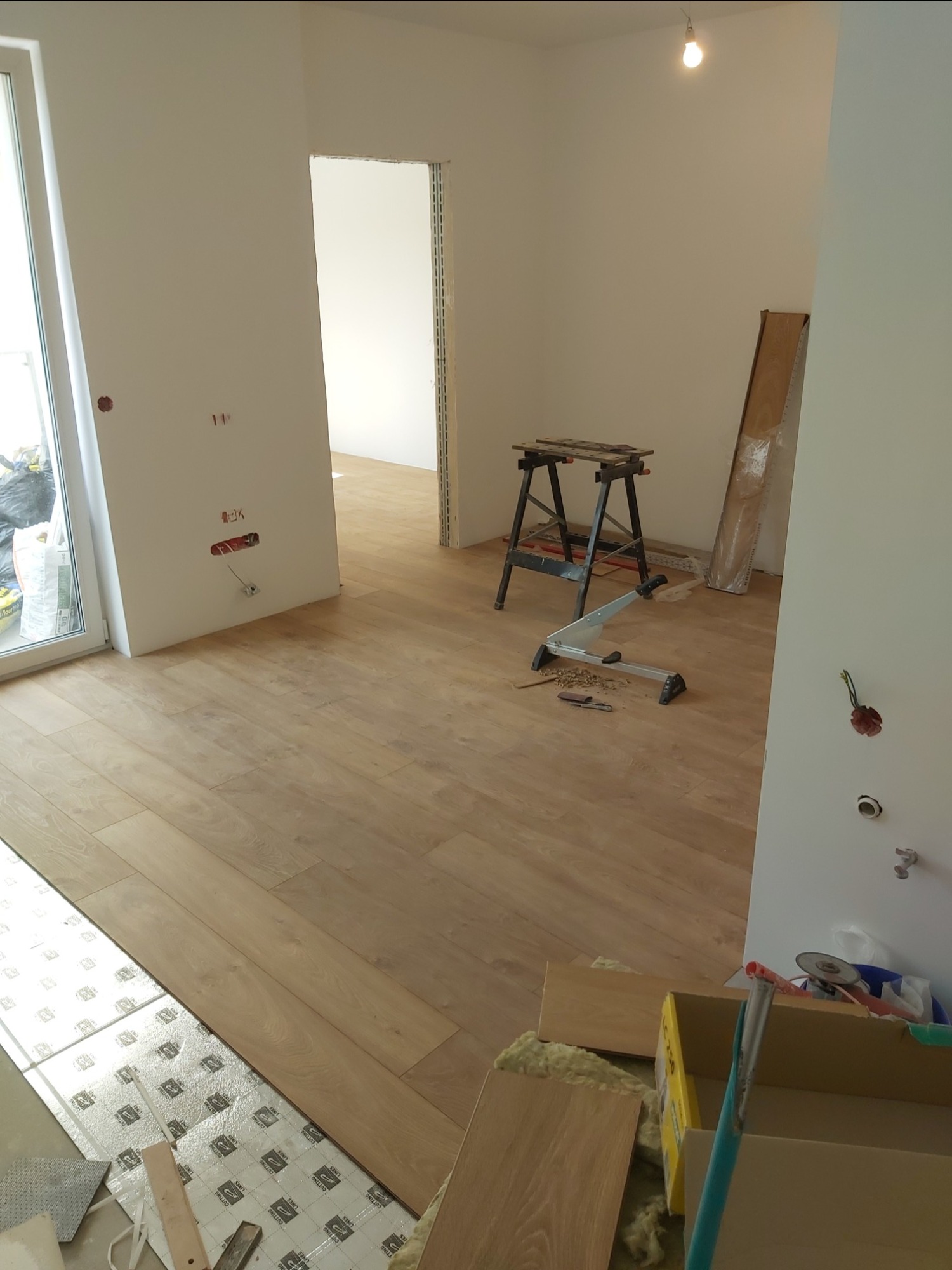 Wnętrze w trakcie układania jasnego parkietu, widoczne narzędzia, podkład, kartony i listwa przypodłogowa, surowy stan ścian z wystającymi kablami elektrycznymi, brak umeblowania.