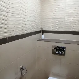 Beżowa łazienka z dekoracyjnymi płytkami ściennymi w fale, chromowanym uchwytem na papier toaletowy i przyciskiem spłuczki, oraz białym dyfuzorem zapachowym na półce.