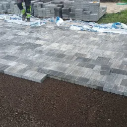 Ułożona szara kostka brukowa na podsypce, z widocznymi zapasami kostki i narzędziami pracy, częściowo przykryta folią ochronną.