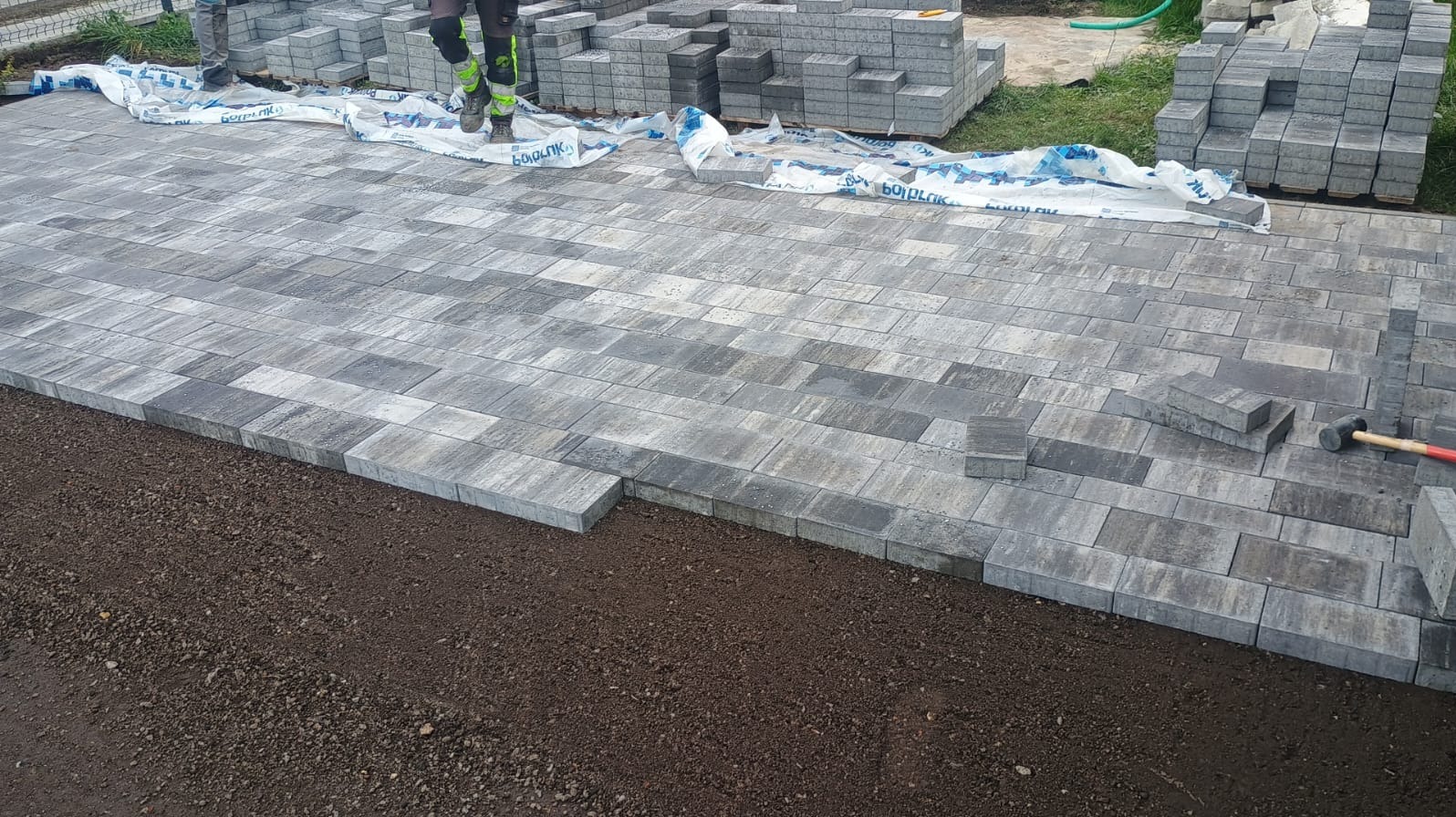Ułożona szara kostka brukowa na podsypce, z widocznymi zapasami kostki i narzędziami pracy, częściowo przykryta folią ochronną.