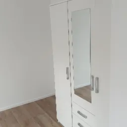 Nowoczesna, biała szafa z lustrem i trzema szufladami, ustawiona na panelach podłogowych w jasnym, minimalistycznym wnętrzu. Widok z góry.