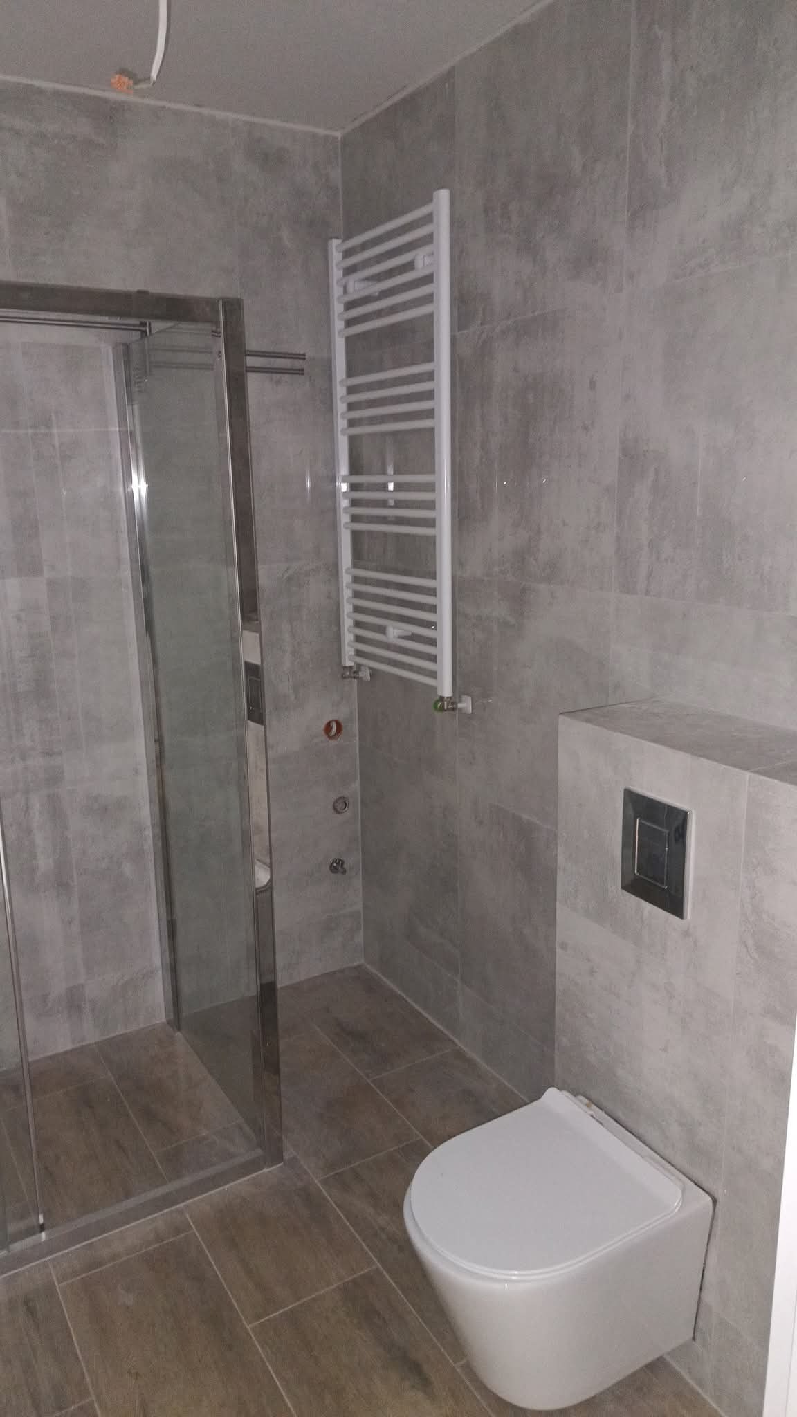 Wykończona łazienka z kabiną prysznicową ze srebrnymi ramami, grzejnikiem drabinkowym i podwieszaną toaletą w szarościach. Płytki imitujące drewno na podłodze.