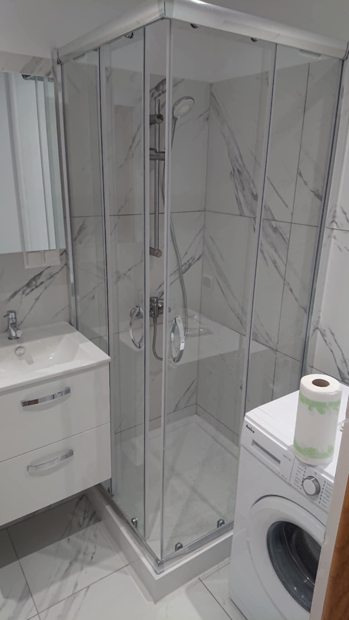 Biała łazienka z kabiną prysznicową narożną, umywalką z szafką i pralką Amica. Marmurowe płytki na ścianach i podłodze. Nowoczesny design.