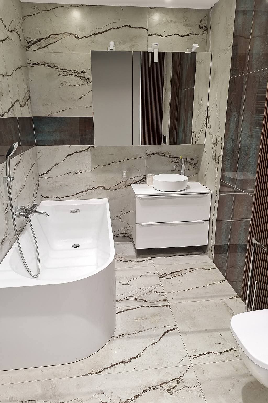 Nowoczesna łazienka z marmurowymi płytkami, białą wanną i szafką pod umywalkę. Minimalistyczne oświetlenie i lustro dodają elegancji. Widoczny fragment toalety.