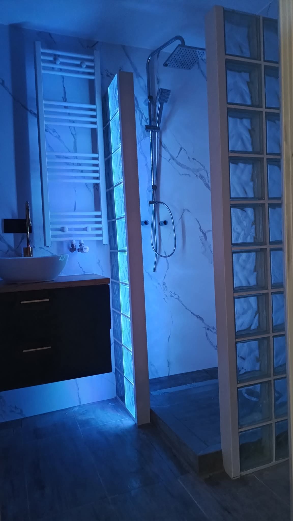Nowoczesna łazienka z kabiną prysznicową oddzieloną ścianką z luksferów, podświetlona niebieskim światłem LED, umywalka na szafce i grzejnik drabinkowy.