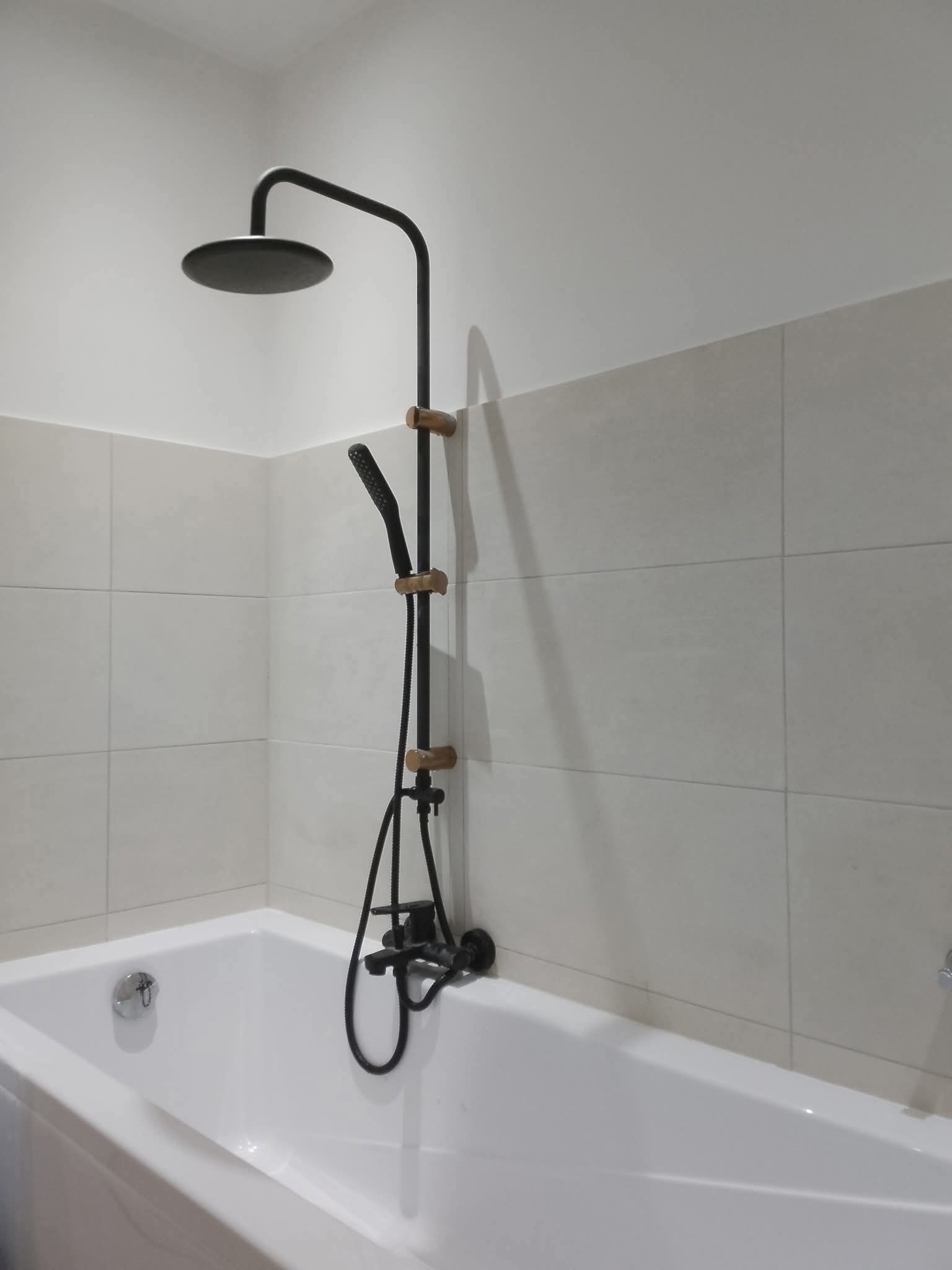 Nowoczesna łazienka: biała wanna z czarną armaturą prysznicową i jasnymi, kwadratowymi płytkami na ścianie. Minimalistyczny design i stonowana kolorystyka.