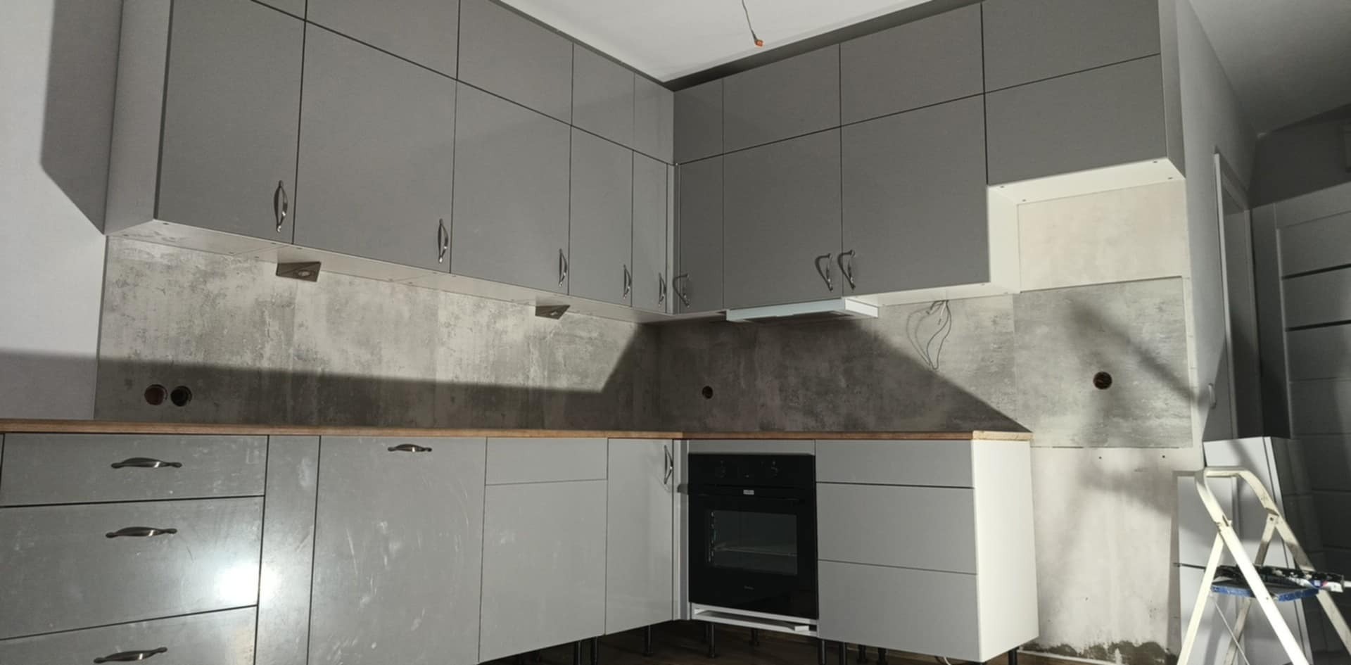 Szara zabudowa kuchenna z płytkami imitującymi beton, widoczne otwory na gniazdka, drabina malarska w rogu. Kuchnia w trakcie montażu, surowy stan.