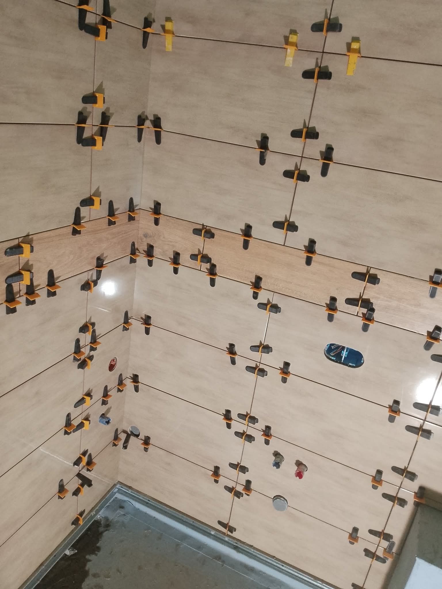 Układanie beżowych płytek w łazience z użyciem systemu poziomowania. Widoczne krzyżyki i kliny. Fragment odpływu liniowego i otwór na instalację elektryczną.