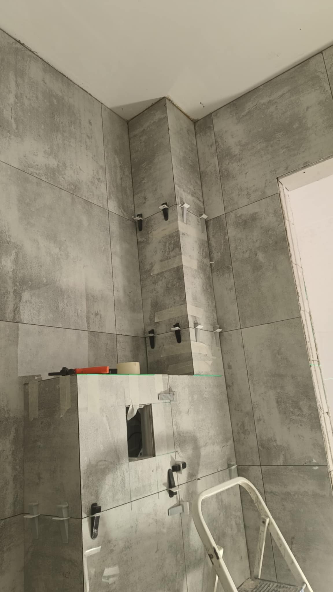 Ściany łazienki w trakcie wykańczania szarymi płytkami imitującymi beton, z użyciem klipsów poziomujących i widoczną drabinką malarską.