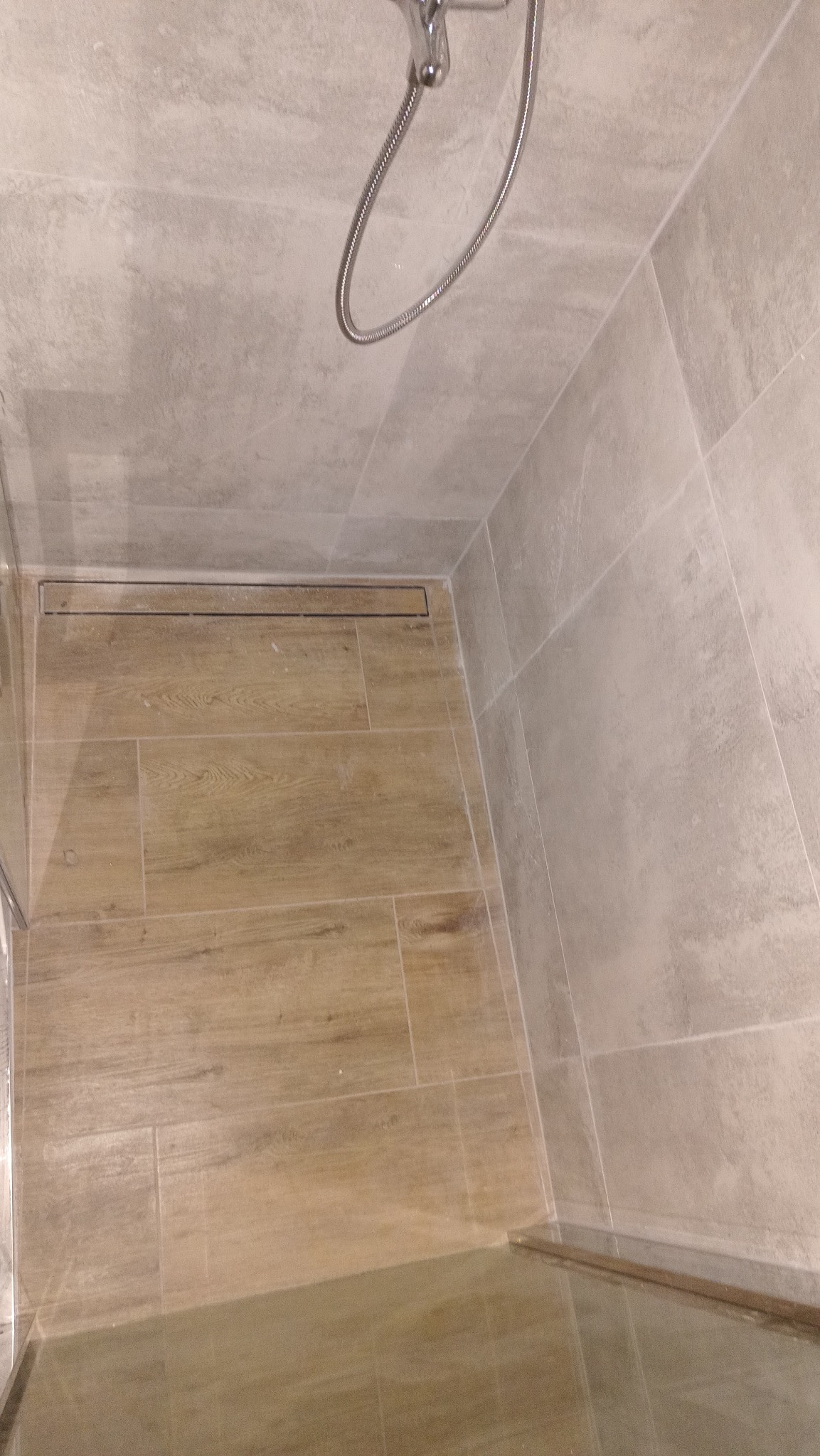 Nowoczesna kabina prysznicowa z odpływem liniowym i płytkami imitującymi drewno na podłodze. Ściany wyłożone jasnymi, beżowymi płytkami.