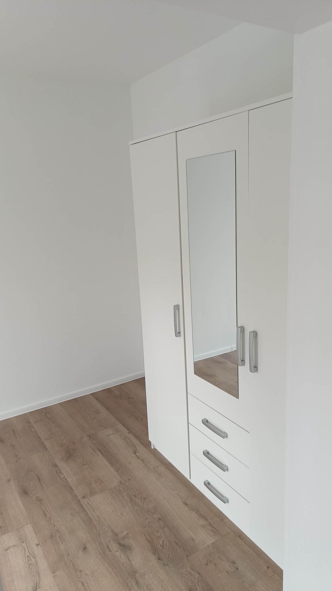 Nowoczesna, biała szafa z lustrem i trzema szufladami, ustawiona na panelach podłogowych w jasnym, minimalistycznym wnętrzu. Widok z góry.