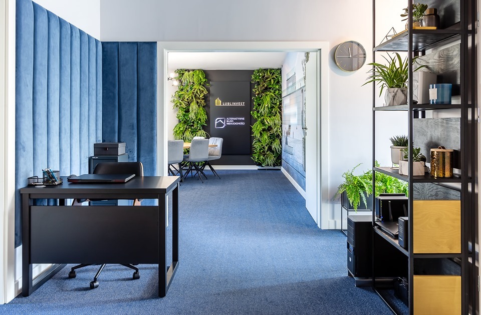 Biuro z niebieską wykładziną i ścianą z zielenią w tle, logo firmy Lublinvest widoczne na czarnej ścianie, nowoczesny design.
