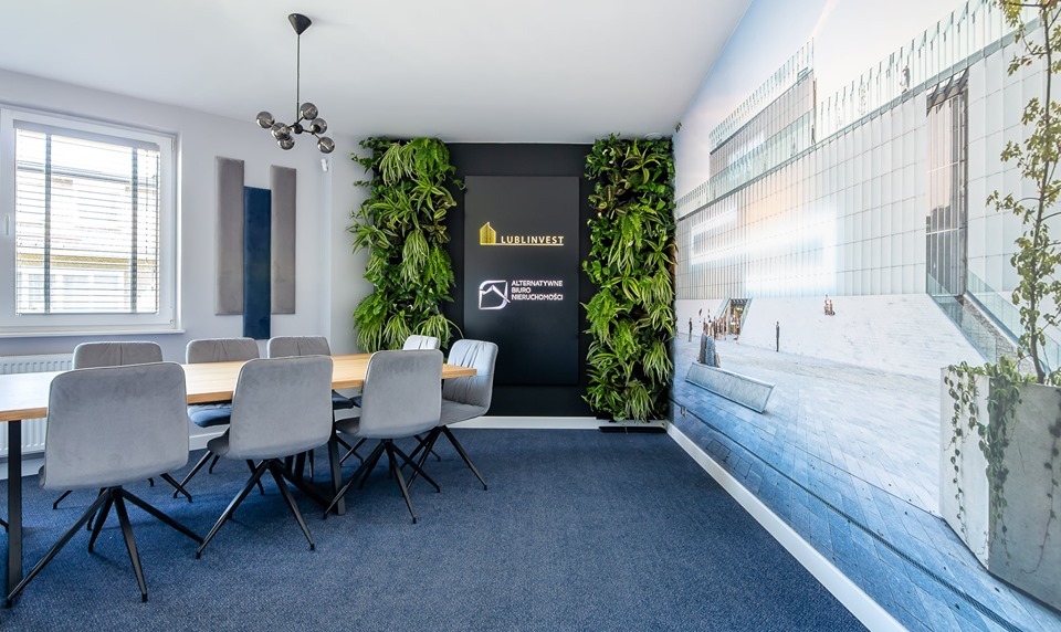 Sala konferencyjna z drewnianym stołem, szarymi krzesłami na kółkach, pionową zielenią na ścianie i tapetą przedstawiającą nowoczesny budynek, logo firmy Lublinvest.