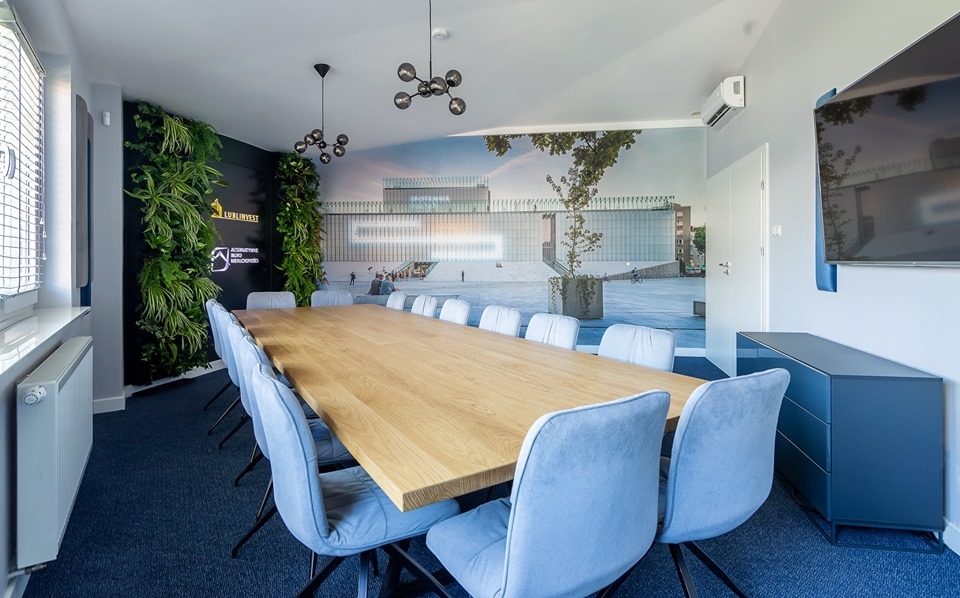 Nowoczesna sala konferencyjna z długim drewnianym stołem, niebieskimi krzesłami, pionowym ogrodem i fototapetą przedstawiającą budynek Muzeum Narodowego w Lublinie.