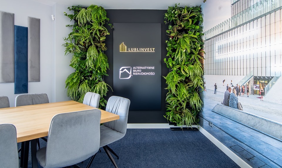 Sala konferencyjna biura nieruchomości z drewnianym stołem, szarymi krzesłami, pionowymi ogrodami z roślin po bokach czarnego panelu z logo firmy Lublinvest i Alternatywne Biuro Nieruchomości...