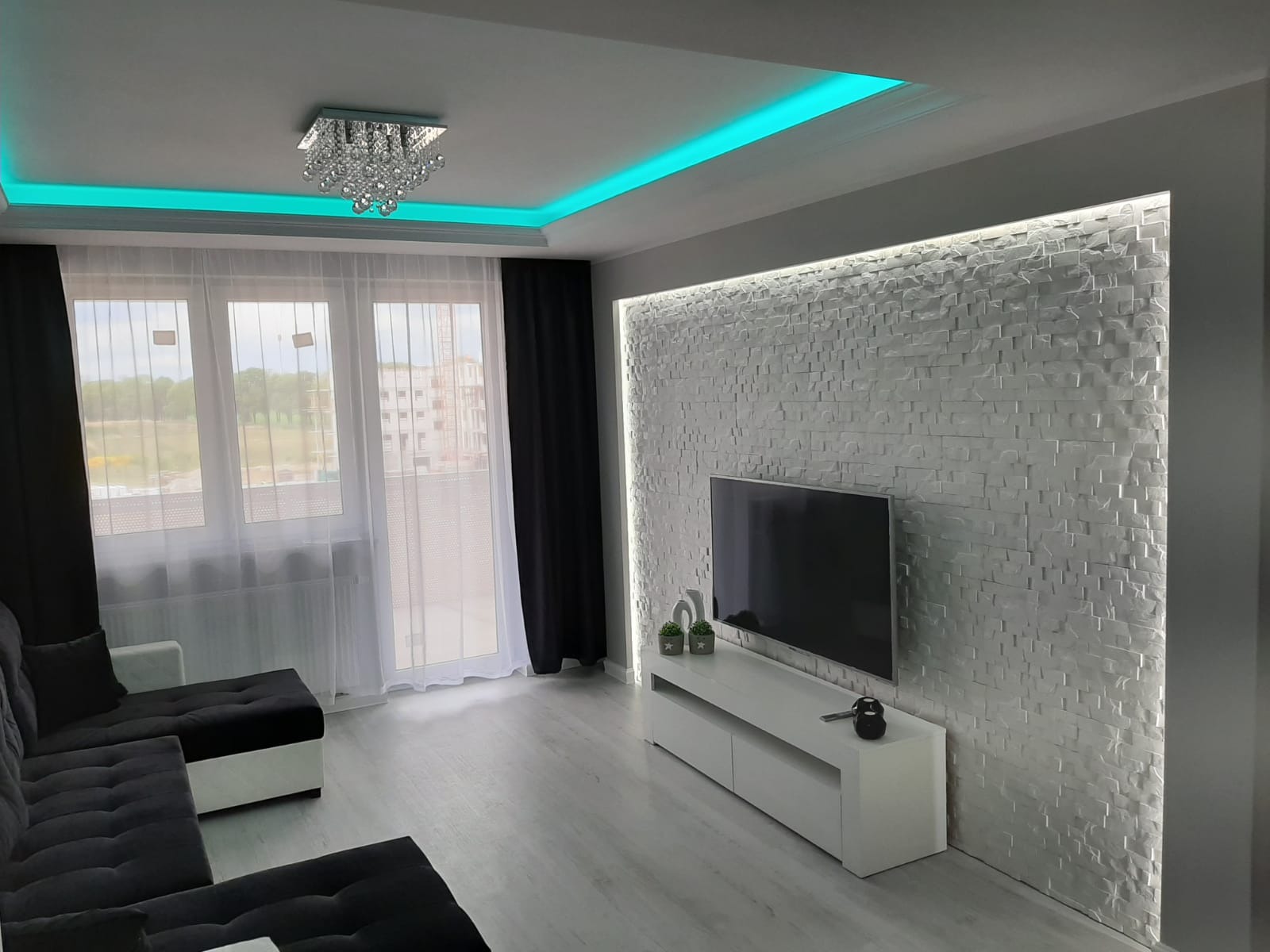 Nowoczesny salon z szarą sofą, białymi meblami RTV i ścianą wyłożoną dekoracyjną, podświetlaną białą cegłą. Sufit z oświetleniem LED w kolorze turkusowym i kryształowy żyrandol.