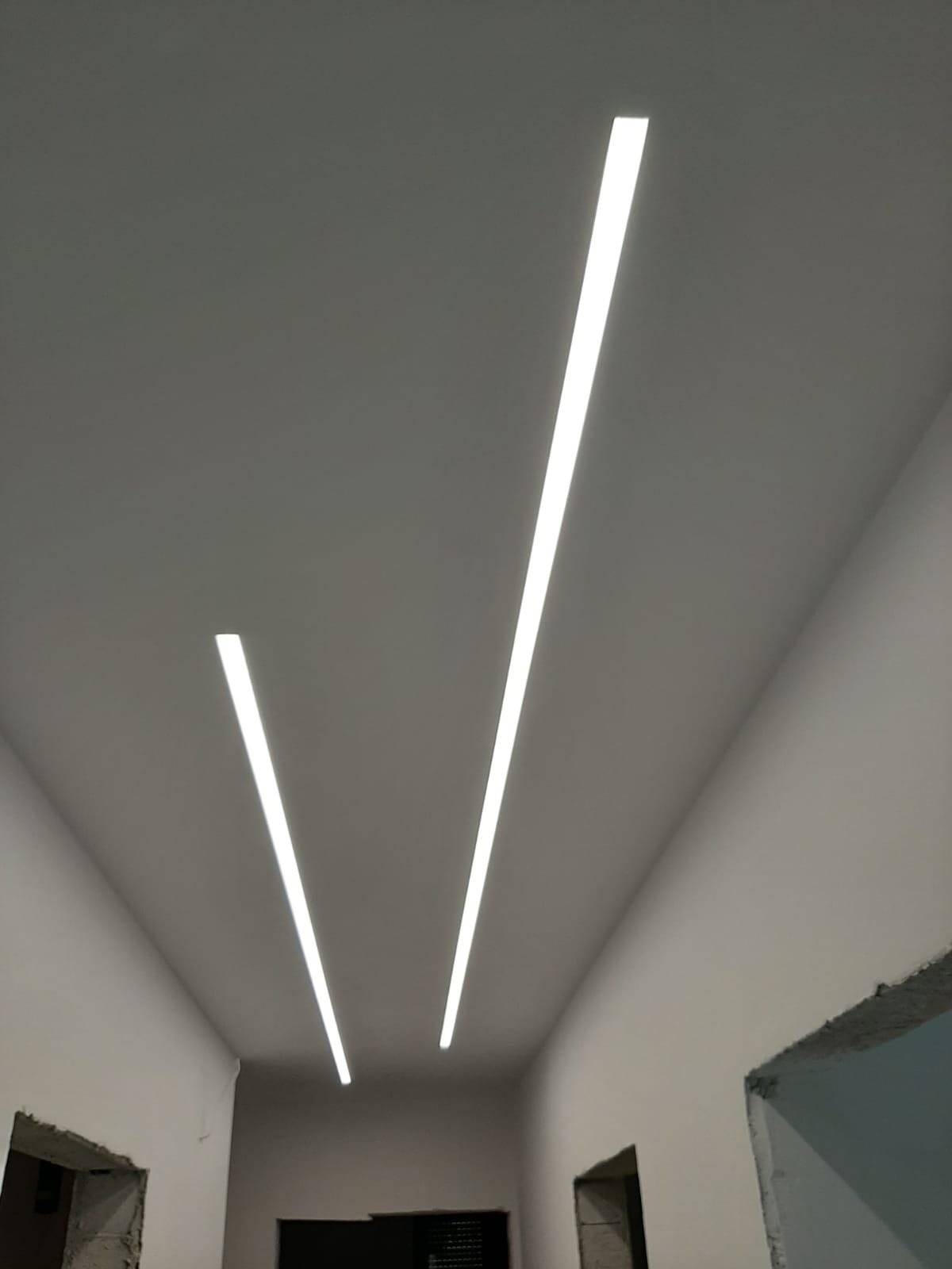 Dwa równoległe pasy LED wpuszczone w sufit, oświetlające korytarz w trakcie prac wykończeniowych, widoczne surowe ościeżnice drzwiowe.