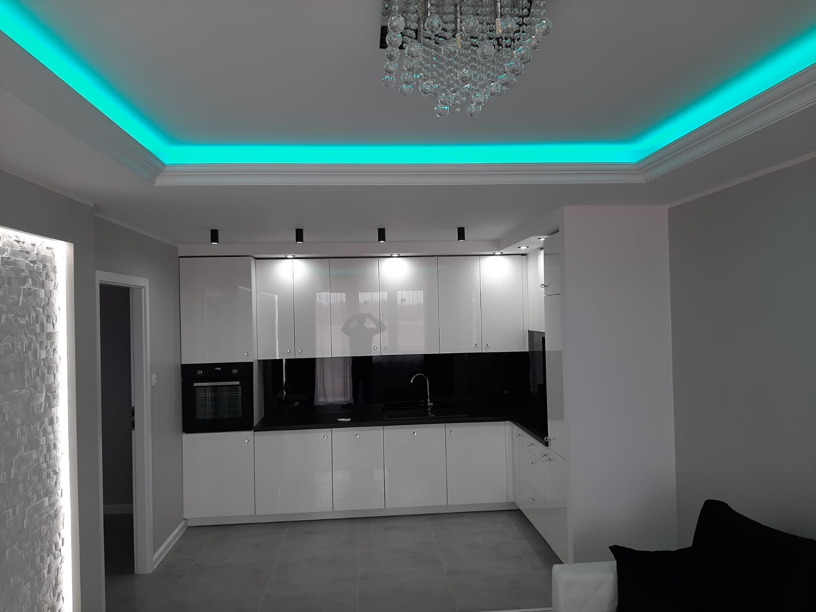 Nowoczesna kuchnia z białymi, lakierowanymi szafkami, czarnym blatem i podświetleniem LED w kolorze turkusowym w suficie oraz dekoracyjnym oświetleniem ściany imitującym kamień.