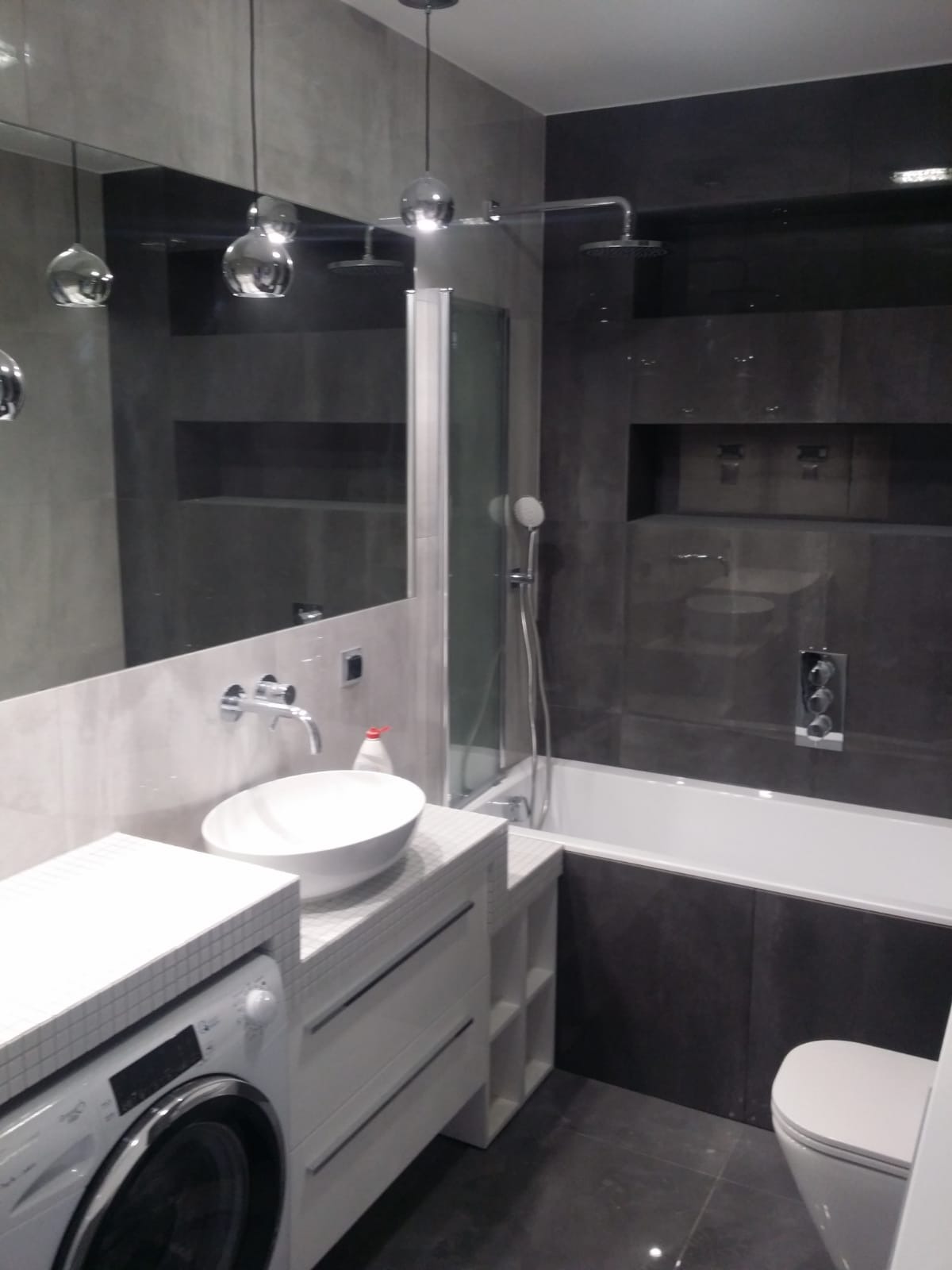 Nowoczesna łazienka z szarymi płytkami, białą umywalką nablatową, pralką pod blatem i kabiną prysznicową za szklaną ścianką, oświetlona wiszącymi lampami z chromowanymi kulami.