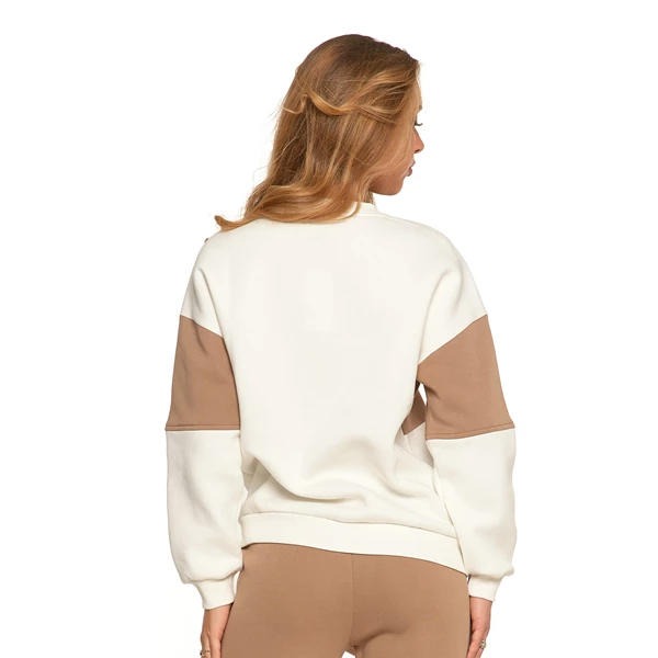 Kobieta tyłem w beżowo-kremowej bluzie z długim rękawem. Włosy blond spięte z tyłu głowy. Gładkie tło, neutralne oświetlenie.