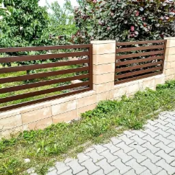 Nowoczesne, brązowe ogrodzenie z poziomych metalowych profili, osadzone na słupkach z jasnych, kwadratowych płytek, widok z poziomu chodnika z kostki brukowej, w tle zieleń ogrodu.