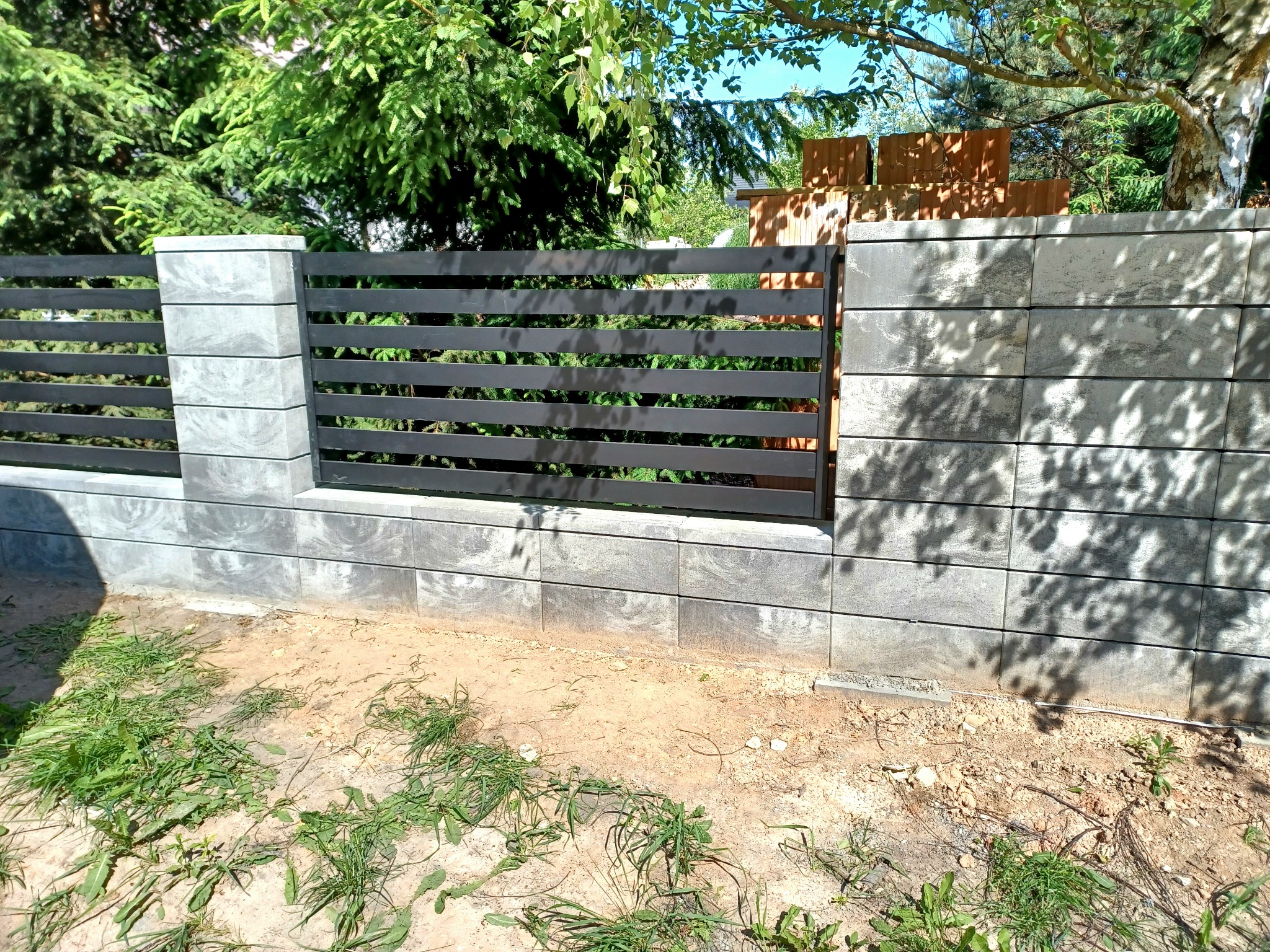 Nowoczesne ogrodzenie z szarych betonowych bloczków i poziomych, czarnych metalowych przęseł, widoczne na tle zieleni drzew i częściowo ułożonych cegieł.