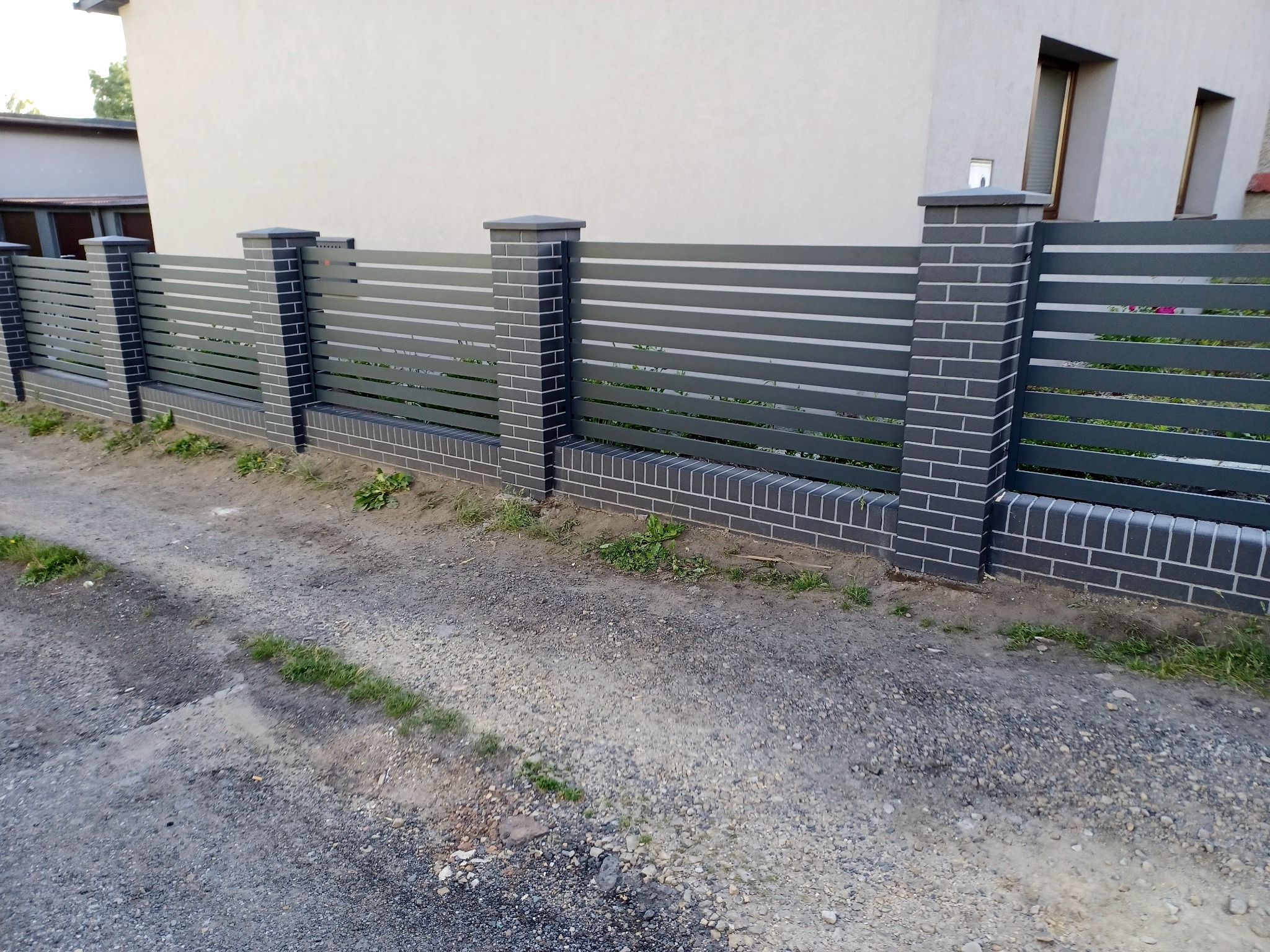 Nowoczesne ogrodzenie panelowe w kolorze grafitowym z ceglanymi słupkami i podmurówką, widok z zewnątrz na szutrową drogę.