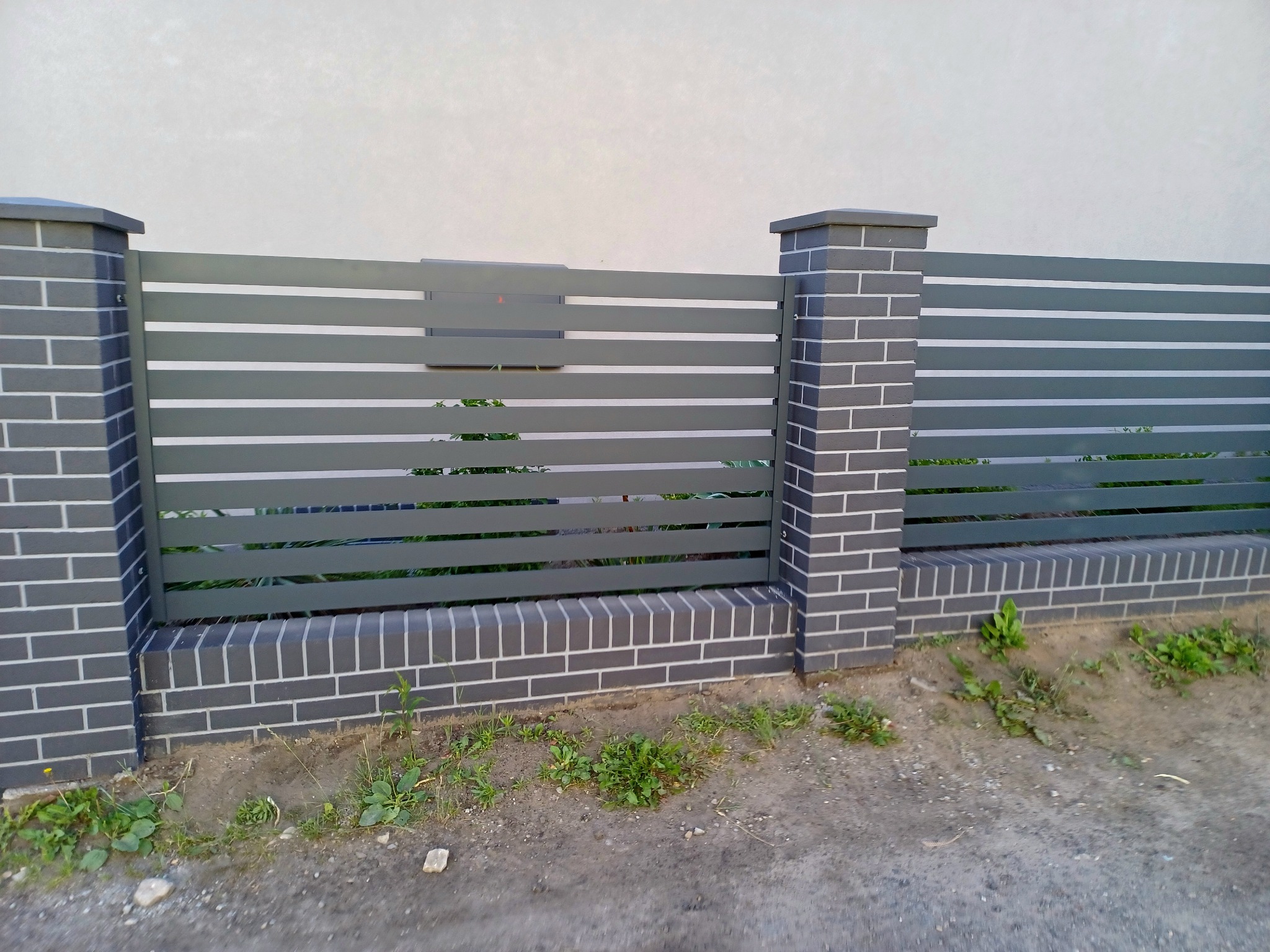 Nowoczesne ogrodzenie z szarych, poziomych paneli metalowych i słupków z szarej cegły klinkierowej z białymi fugami.
