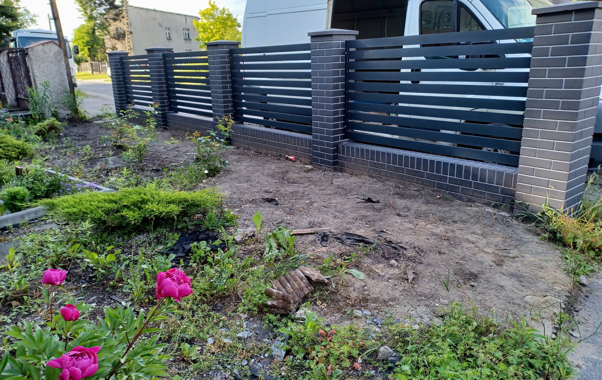 Nowoczesne ogrodzenie z szarej cegły i poziomych metalowych paneli, widoczne na tle zieleni i zaparkowanego busa.