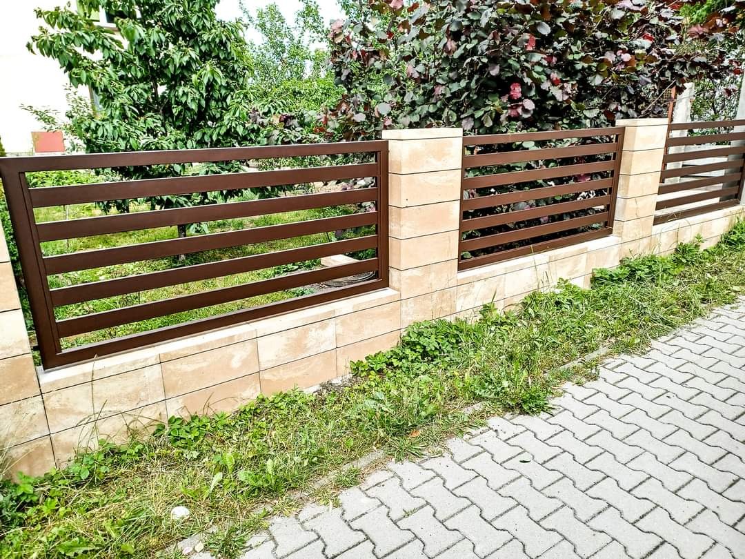 Nowoczesne, brązowe ogrodzenie z poziomych metalowych profili, osadzone na słupkach z jasnych, kwadratowych płytek, widok z poziomu chodnika z kostki brukowej, w tle zieleń ogrodu.