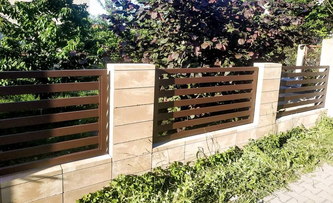 Nowoczesne ogrodzenie z poziomych, brązowych profili metalowych, osadzone na niskim murku z beżowych, betonowych bloczków, otoczone zielenią.